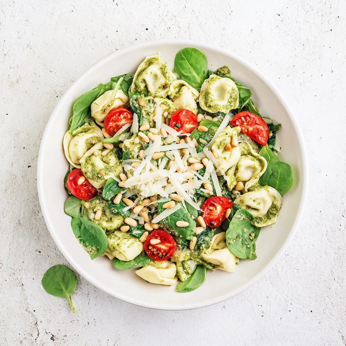 A close-up of chilled pesto tortellini salad, featuring bright cherry tomatoes and fresh spinach leaves for a refreshing summer meal.  