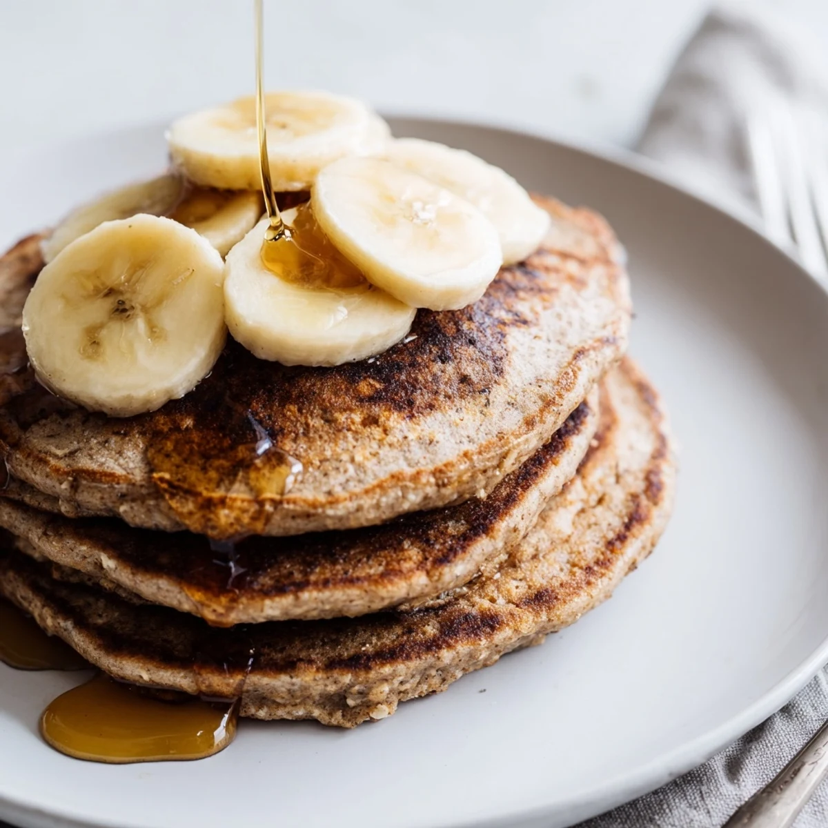 Stack of fluffy Banana Oat Pancakes served on a white plate with a side of yogurt and berries, a perfect easy brunch recipe.