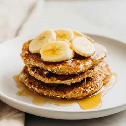 Golden-brown Banana Oat Pancakes sizzling in a skillet, topped with fresh banana slices and a drizzle of maple syrup for a wholesome breakfast.