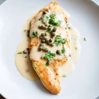 Golden, pan-fried Creamy Chicken Piccata breasts simmering in a bright lemon and caper sauce, garnished with fresh parsley.